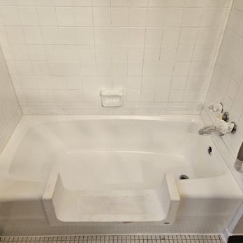 Bath Magic - A white bathtub is installed in a bathroom with white tiled walls. The tub appears to be in the process of repair or renovation, with exposed plumbing fixtures and a plastic bag covering part of the pipes. The surrounding tiles are uniformly white, and the floor features small square tiles. The image suggests ongoing work on the bathtub or plumbing system. - Refinishing Services Near Me