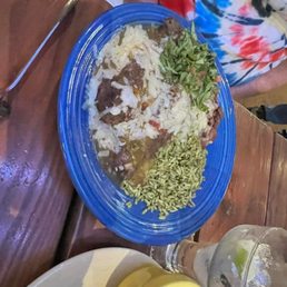 Blue Corn Chile Rellenos - A meal served on a blue plastic plate includes beef with sauce, white rice, shredded lettuce, and melted cheese. A small bowl of salsa and a glass of water with lime are also visible on the wooden table. The setting appears casual, with a person wearing a colorful tie-dye shirt nearby.