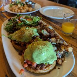 Chorizo Benedict and cheese curds - A white oval plate holds two eggs Benedict topped with green avocado slices and a sprinkle of spices. The eggs are served on toasted English muffins with a meat filling, likely sausage or bacon. To the side, there are crispy breakfast potatoes with some chopped herbs or vegetables. A small side salad with mixed greens and cherry tomatoes is also present. The plate is on a wooden table, with a glass of orange juice, an empty plate, and a water bottle in the background.