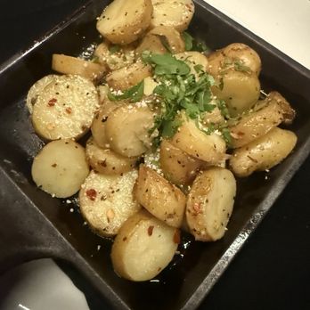 Sinigual - The image shows a black rectangular dish filled with roasted potato pieces, seasoned with herbs, spices, and possibly garlic. The potatoes are garnished with chopped cilantro and sprinkled with coarse salt and red pepper flakes. The dish is placed on a white surface with a black napkin partially visible underneath. - Mexican Near Me - Manhattan, New York