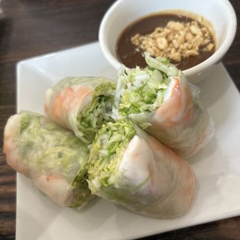 Pho Saigon - A1. Spring Roll - Four Vietnamese spring rolls filled with shredded lettuce and vegetables are arranged on a white square plate. A small bowl of dipping sauce with chopped peanuts or crushed ingredients is placed beside the rolls. The dish is set on a dark wooden table, with part of another plate visible in the background. - Soup Near Me - Dover, Delaware