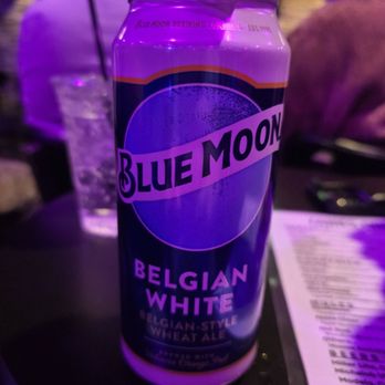 Zanies - A can of Blue Moon Belgian White beer, a Belgian-style wheat ale, is placed on a table with a slice of watermelon on top. The background shows blurred figures and a menu, indicating a social setting such as a bar or restaurant. The lighting has a purple hue, typical of nightlife venues. - Comedy Clubs Near Me - Rosemont, Illinois