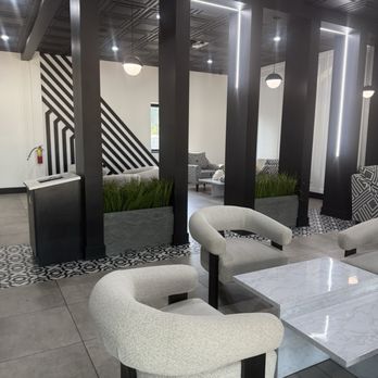 Sada Coffee - The image shows a modern interior space with black structural columns and a ceiling with a geometric pattern. The area includes seating with white upholstered chairs and marble-topped tables. Decorative planters with green plants are positioned between the columns, and the background features patterned walls and additional seating. The lighting includes pendant lights and LED accents, creating a sleek, contemporary atmosphere. - Desserts Near Me - Palos Hills, Illinois