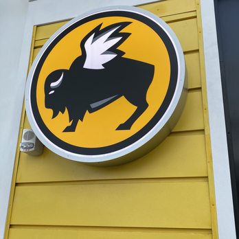 Buffalo Wild Wings - The image shows a circular sign with a black creature that has wings, a beak, and a tail, set against a yellow background. The sign is mounted on a yellow wall with horizontal siding, and a small fire alarm or sensor is visible below the sign. The sign appears to be part of a business or establishment's branding or logo. - Chicken Wings Near Me - Warwick, Rhode Island