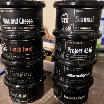 14er Boulder - Great strains! - Multiple black containers with strain names and logos, including 'Mac and Cheese,' 'Shamash,' and 'Jack Herer,' are stacked on a wooden surface. - Vape Shops Near Me - Boulder, Colorado