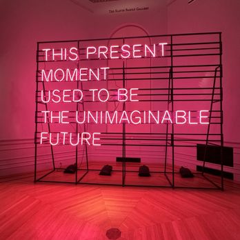 Renwick Gallery - Pink neon text in an art gallery reads: 'This present moment used to be the unimaginable future.' The sign is mounted on a black metal frame with a wooden floor and ornate ceiling details. - Art Galleries Near Me - Washington, DC, District of Columbia