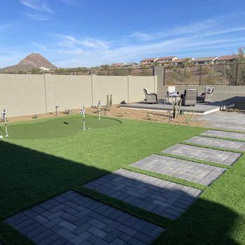 Diamond Cut Landscaping - The image shows a backyard with a well-maintained artificial grass putting green, marked with flags and golf balls. Adjacent to the green is a paved pathway leading to a patio area with outdoor chairs and a table. The yard is enclosed by a concrete block wall, with a mountain and residential houses visible in the distance under a clear blue sky. - Landscaping Near Me - Phoenix, Arizona