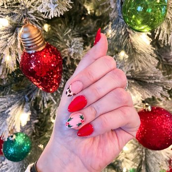 Nail's Corner - December set- made my vision come alive and loved them! - A person's hand with decorated nails is positioned in front of a Christmas tree adorned with ornaments, including red, green, and glittery baubles. The nails are painted in holiday-themed designs, featuring red polish, holly leaves, and a leopard print accent. The tree has white-tipped branches and is illuminated with small lights, creating a festive holiday scene. - Nail Technicians Near Me - Keizer, Oregon