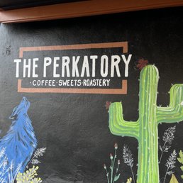 a sign for a coffee shop - A wall features a painted sign for 'The Perkatory,' a coffee shop, with artwork including a blue bird and a green cactus, on a dark background.