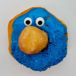 Doughnuts - A donut features blue icing with candy eyes and a large, rounded, yellowish nose made of icing or fondant, resembling Grover from Sesame Street. The donut has blue sprinkles on part of the icing and is set against a plain white background.