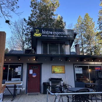 Bistro Espresso - The image depicts the exterior of Bistro Espresso, a small restaurant with outdoor seating arrangements including tables and chairs. A sign on the building indicates additional seating is available in the backyard. The building has a dark-colored facade with a red door and a window. There are string lights hanging above the outdoor area, and a sign with the business name and offerings is visible at the top. The background includes tall trees and a clear blue sky. - Coffee & Tea Near Me - Arnold, California