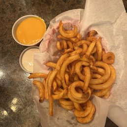 Curly fries - A basket of curly fries is placed on a dark, textured table, lined with white parchment paper. Two small cups of dipping sauces, one yellow (likely cheese or mustard) and one white (possibly ranch or mayonnaise), are positioned nearby. The fries are golden-brown and appear crispy, with some curled and twisted in shape.
