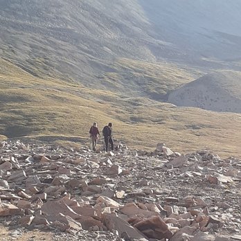 Mount Sherman - Two hikers are walking on a rocky trail in a mountainous area with grassy slopes and distant peaks. - Hiking Near Me - Fairplay, Colorado