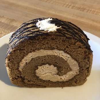 Belle's Bread - Coffee roll cake slice - A slice of chocolate roll cake features a spiral pattern of chocolate sponge and cream filling. The cake is topped with a small dollop of cream and a drizzle of chocolate sauce. It is placed on a white plate, with a wooden surface in the background. The cake appears moist and well-made, with a rich chocolate appearance. - Bakeries Near Me - Columbus, Ohio