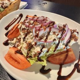 Wedge salad - A wedge salad served on a white oval plate includes chopped iceberg lettuce, tomato wedges, and thinly sliced red onions. The salad is garnished with crumbled cheese and drizzled with a dark balsamic glaze and additional dressing. The presentation is simple, with the ingredients arranged neatly on the plate, and the salad appears fresh and appetizing.