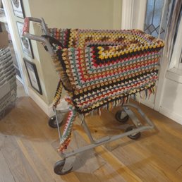 Crocheted shopping cart - A metal shopping cart with a multicolored crocheted cover is positioned indoors near a wall and window, in a retail setting.