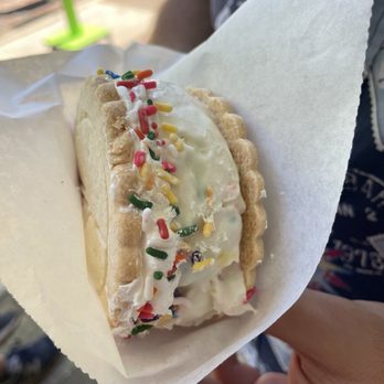 Peace Pie - Birthday Cake Ice Cream Sandwich - A close-up of a birthday cake ice cream sandwich featuring vanilla ice cream with colorful sprinkles on top, sandwiched between two cookie-like wafers. The ice cream is slightly melting, and the sandwich is wrapped in white paper, held by a person's hand. The background shows an outdoor setting with blurred elements, including a person and a green object. - Desserts Near Me - St. Augustine, Florida