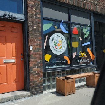 Rooted MKE - The storefront window displays colorful abstract shapes and the Rooted MKE logo, which includes the text 'Learn With Us.' The window also has the word 'Bookstore' written on it. Two wooden planters are positioned in front of the window, and the building has a brick exterior with an orange door to the left. The window design suggests a focus on education and community engagement. - Bookstores Near Me - Milwaukee, Wisconsin