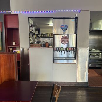 Project Burger - Local sodas - A soda fountain station featuring multiple drink dispensers is positioned against a wall in a restaurant setting. The dispensers are labeled and arranged in a row, with a stainless steel panel behind them. The background shows kitchen equipment and shelves with supplies, indicating an open kitchen layout. The area is part of a casual dining environment with wooden and metal furnishings. - American Near Me - Alameda, California