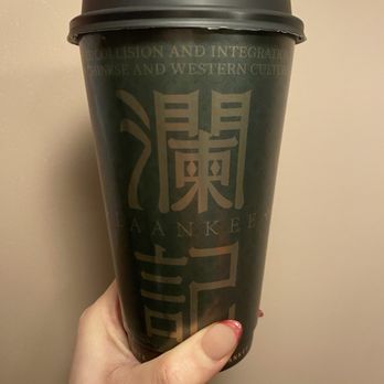 Laankee - Yashi milk tea -IG: susanlamx - A person holds a black paper cup with a plastic lid, featuring Asian characters and the word 'Yankee' printed on it, against a plain wall. - Bubble Tea Near Me - Brooklyn, New York