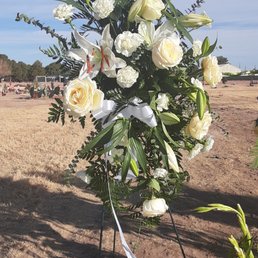 A floral arrangement with white roses, lilies, and various green foliage is displayed on a stand outdoors. The background shows a field with people and structures, under a partly cloudy sky. The arrangement includes a white ribbon tied to the stand, suggesting it may be for a memorial or special event.