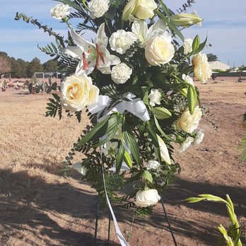 Debbie's Bloomers - A floral arrangement with white roses, lilies, and various green foliage is displayed on a stand outdoors. The background shows a field with people and structures, under a partly cloudy sky. The arrangement includes a white ribbon tied to the stand, suggesting it may be for a memorial or special event. - Florists Near Me - El Paso, Texas