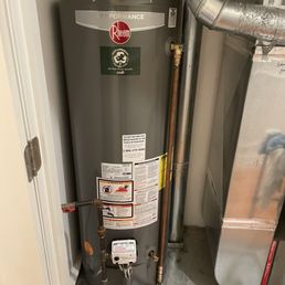 New hot water heater - A Rheem hot water heater is positioned in a small utility space, connected to copper and metal pipes, with safety and instruction labels visible.