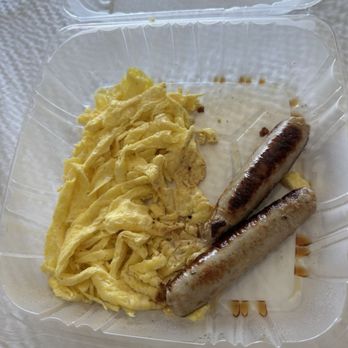 Elmer's Restaurant - Chicken Zenner's Chicken Apple Sausage Links & Eggs - The image shows a clear plastic takeout container filled with scrambled eggs and two grilled sausage links. The eggs are yellow and slightly fluffy, while the sausages are browned with grill marks. The container is placed on a textured white surface, and the lid is partially visible at the top of the image. - Diners Near Me - Eugene, Oregon