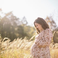 Ashlee Wilcox Photography - Maternity photo. Expectant mother to be stands in a golden field of grass, holding wildflowers and smiling down at her baby bump.