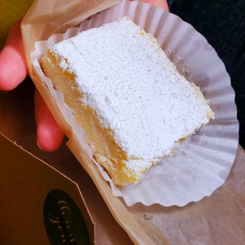 Suger Bōle - Lemon bar - A square lemon bar with a dusting of powdered sugar on top is held in a person's hand. The dessert is placed in a white paper cupcake liner and partially wrapped in brown parchment paper. The background includes a cardboard box with a black sticker and a wooden surface. - Beer, Wine & Spirits Near Me - Biddeford, Maine