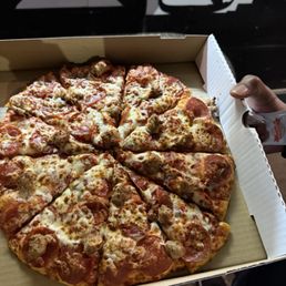 Medium pepperoni and sausage pizza - A pizza with slices of pepperoni and sausage is displayed in an open cardboard pizza box. The pizza has a cheese topping and is cut into slices. The person holding the box is visible, with their hand gripping the side. The background includes a black and white wall with silhouettes, and the person's feet are visible at the bottom of the image.