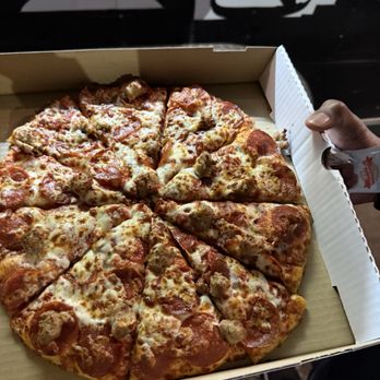Pizza Leon - Medium pepperoni and sausage pizza - A pizza with slices of pepperoni and sausage is displayed in an open cardboard pizza box. The pizza has a cheese topping and is cut into slices. The person holding the box is visible, with their hand gripping the side. The background includes a black and white wall with silhouettes, and the person's feet are visible at the bottom of the image. - Pizza Near Me - Austin, Texas