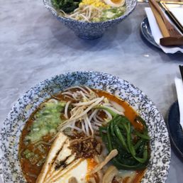 Two bowls of ramen with toppings like boiled eggs, vegetables, and meat are placed on a marble table at a restaurant, with chopsticks and utensils nearby.
