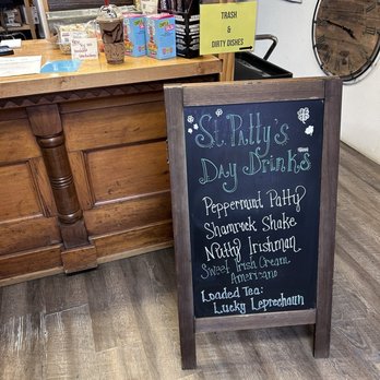 Allsorts - a chalkboard sign in a store - A chalkboard sign in a shop or cafe lists St. Patty's Day themed drinks such as Peppermint Patty, Shamrock Shake, Nutty Irishman, and Lucky Leprechaun. The sign is positioned on a wooden floor near a wooden counter with various items and a yellow sign indicating trash and dirty dishes. The background includes a large clock on the wall and some electrical panels, suggesting an indoor retail or cafe environment. - Antiques Near Me - Sutherland, Nebraska