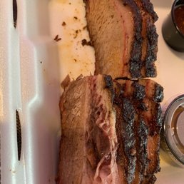 Brisket - Two slices of smoked brisket with a dark, crispy bark are placed in a foam takeout container, with some juices and seasoning visible.
