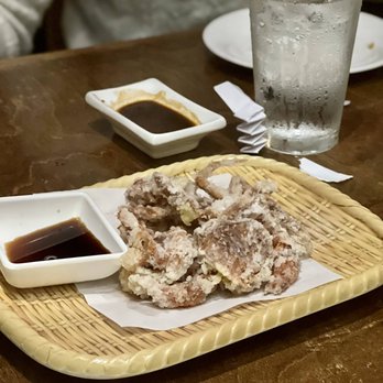 Yuzu Sushi - soft shell crab - Fried soft shell crab is presented on a woven tray lined with paper, accompanied by a small dish of soy-based dipping sauce. The tray is placed on a wooden table, with a glass of water and a small dish of soy sauce visible in the background. The setting appears to be a restaurant dining area. - Japanese Near Me - Chamblee, Georgia