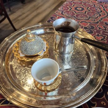 Cafe Baladi - Turkish coffee - A round silver tray features a Turkish coffee pot, a small white cup with a handle, and a decorative container with a lid, placed on a table with a patterned tablecloth. The background shows chairs and a wooden floor, indicating a restaurant setting. - Lebanese Near Me - Malvern, Pennsylvania