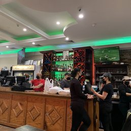 Bar area - The image depicts the bar area of a restaurant, featuring staff members wearing masks working behind a wooden counter. There are multiple television screens mounted on the wall, displaying sports and other content. The bar has a display of various bottles of alcohol, and the ceiling has recessed lighting with green accent lights. Several customers or staff are engaged in activities at the counter, and the overall setting appears to be a casual dining or sports bar environment.