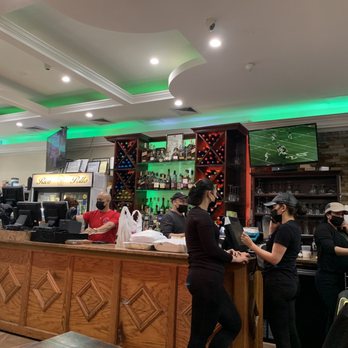 Rico Pollo - Bar area - The image depicts the bar area of a restaurant, featuring staff members wearing masks working behind a wooden counter. There are multiple television screens mounted on the wall, displaying sports and other content. The bar has a display of various bottles of alcohol, and the ceiling has recessed lighting with green accent lights. Several customers or staff are engaged in activities at the counter, and the overall setting appears to be a casual dining or sports bar environment. - Spanish Near Me - Brooklyn, New York
