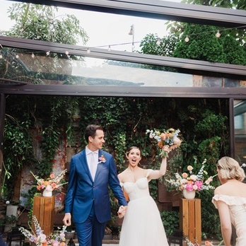 MyMoon - Ceremony deck - A bride and groom are standing together indoors, holding hands and smiling. The bride is wearing a white wedding dress and holding a bouquet, while the groom is dressed in a blue suit. The background features lush greenery, floral arrangements, and decorative lighting, indicating a wedding celebration. A guest is visible on the right side of the image, and the setting includes wooden flooring and floral decor. - Venues & Event Spaces Near Me - Brooklyn, New York