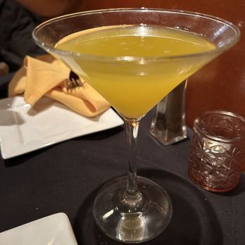 Frix's Fire Grill - Mango martini? With tanteo tequila - A yellow-green cocktail served in a martini glass is placed on a black table, with a napkin, a small glass, and a plate nearby. - Latin American Near Me - Chester, Maryland