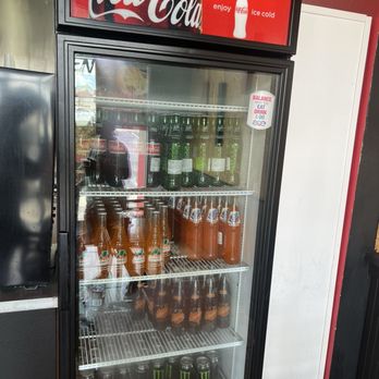 Dogos VIP - A black Coca-Cola refrigerator with a glass door displays bottled drinks including soda, juice, and energy drinks. The top sign features the Coca-Cola logo and the phrase 'enjoy ice cold.' The interior shelves hold bottles of soda, juice, and Monster energy drinks, with some bottles of orange juice and other beverages visible. The refrigerator is situated indoors, next to a white wall and a black surface. - Hot Dogs Near Me - Albuquerque, New Mexico