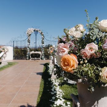 Villa de Amore - Floral team! - Flower arrangements in urns are positioned along a tiled rooftop terrace, with a pathway of flower petals leading to a wedding arch decorated with flowers and greenery. The scene is set outdoors under a clear blue sky, suggesting a wedding or special event setup on a rooftop garden or terrace. - Venues & Event Spaces Near Me - Temecula, California