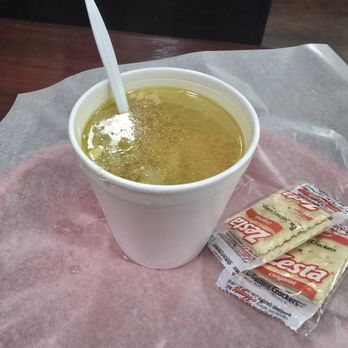 Blaze-N-Grill - A white disposable cup contains hot soup with a clear, yellowish broth. A plastic spoon is inserted into the soup. The cup is placed on a tray lined with pink paper, with two saltine crackers in individual wrappers positioned beside it. The background is dark and out of focus. - Burgers Near Me - Chicago, Illinois