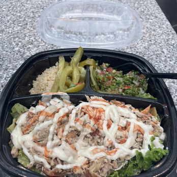Pita Thyme - Chicken shawarma plate on lettuce with quinoa, tabbouleh and pickles - A black plastic takeout container on a granite table contains chicken shawarma topped with mayonnaise, a side of tabbouleh salad with chopped vegetables, and pickled peppers. The container is open, with a black fork inside, and a clear plastic lid is placed behind it. The meal includes traditional Middle Eastern dishes, presented in a segmented container for easy access. - Lebanese Near Me - Boston, Massachusetts