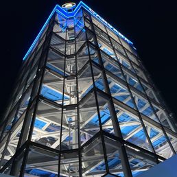 A multi-story car vending machine with transparent glass walls and a steel framework, illuminated by blue neon lights at night. The structure displays several cars on different levels, with visible internal mechanical components. The building's design emphasizes modern architecture and automotive retail innovation.