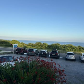 Surfside - View from front deck - A parking lot with several cars is situated near a grassy area with bushes, overlooking the ocean under a clear blue sky. - Seafood Near Me - Montauk, New York