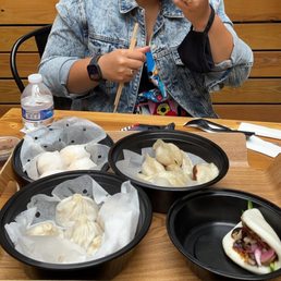 Steam buns, har gow, pork belly buns, and fried dumplings - A person wearing a denim jacket is seated at a wooden table, holding a steamed bun and chopsticks. The table has several black containers filled with different types of dumplings, and a water bottle is visible on the left. The background features wooden paneling, indicating an indoor dining setting.