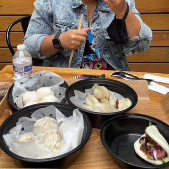 Mr Bao - Steam buns, har gow, pork belly buns, and fried dumplings - A person wearing a denim jacket is seated at a wooden table, holding a steamed bun and chopsticks. The table has several black containers filled with different types of dumplings, and a water bottle is visible on the left. The background features wooden paneling, indicating an indoor dining setting. - Shanghainese Near Me - Brooklyn, New York