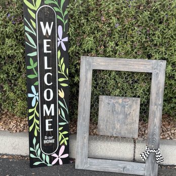 Board & Brush - A painted wooden welcome sign with floral designs leans against a hedge, alongside a wooden frame with chicken wire and a bow. - Signmaking Near Me - Gilbert, Arizona
